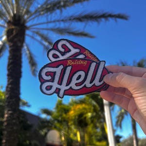 May include: Two red and white embroidered patches with the text "Raising Hell" in white lettering. The patches are held in a hand against a blurred background of palm trees and a blue sky.