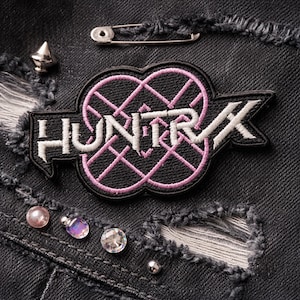 May include: Close-up of a black patch with the word "HUNTRX" in white, outlined in black, and a pink geometric design. The patch is on a distressed black denim surface, with a safety pin and decorative studs.