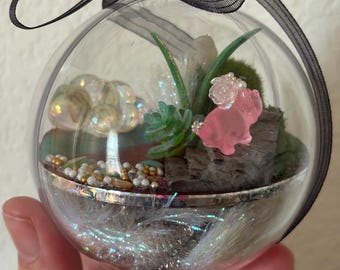 Diorama diminuto de dinosaurio triceratops rosa en un adorno transparente, escena diminuta y brillante de otro mundo con una montaña y nubes, adorno