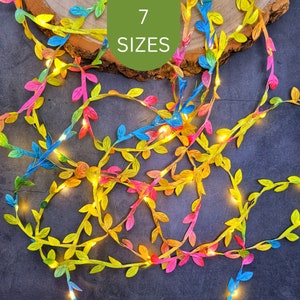 May include: A vibrant leaf garland with integrated fairy lights. The leaves are in shades of yellow, pink, blue, and green. The image includes the text "7 SIZES" in a green circle. The garland is arranged on a dark surface.