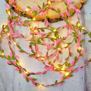 May include: Pink and green leaf garland with warm white LED lights. The garland is wrapped around a wooden slice.