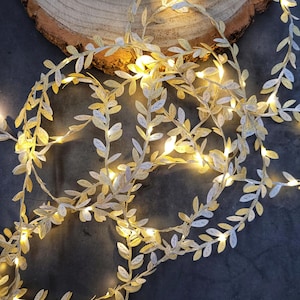 Könnte beinhalten: Eine dekorative Blattgirlande mit kleinen, warmweißen LED-Lichtern. Die Blätter sind gold- und silberfarben und erzeugen einen schimmernden Effekt. Die Girlande ist auf einer dunklen Oberfläche angeordnet, mit einer Holzscheibe im Hintergrund.