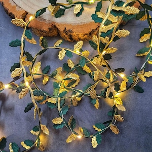 May include: A decorative string of lights featuring alternating dark green and metallic gold holly leaves. The flexible wire is intertwined with small, warm white LED bulbs that emit a soft glow throughout the length of the garland.