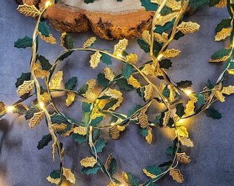May include: A decorative string of lights featuring alternating dark green and metallic gold holly leaves. The flexible wire is intertwined with small, warm white LED bulbs that emit a soft glow throughout the leafy garland.