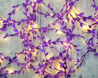 May include: A string of purple artificial vine leaves intertwined with small, warm white LED fairy lights. The flexible wire is covered in numerous small, pointed purple leaves, creating a decorative garland effect with glowing light points.