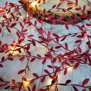 May include: Red leaf garland with warm white fairy lights. The garland is made of fabric leaves and is perfect for adding a touch of festive cheer to any space.
