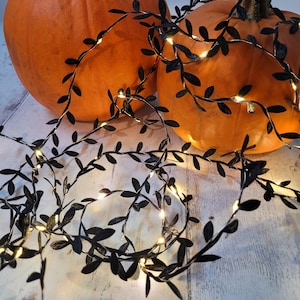 Puede incluir: Dos calabazas naranjas están decoradas con una guirnalda de hojas negras con pequeñas luces blancas. La guirnalda está colocada sobre las calabazas, creando una exhibición festiva de otoño o Halloween. Las calabazas están sobre una superficie de madera clara.
