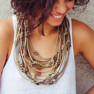 May include: A layered necklace in shades of green and brown, with multiple knotted sections. The necklace is made of cord. The woman is wearing a white vest top.