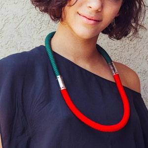 Puede incluir: Una mujer con el pelo corto y castaño oscuro lleva una blusa negra y un collar rojo y verde. El collar está hecho de cuerda gruesa y tiene un acento plateado.