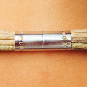 May include: Close-up of a necklace with a silver clasp and beige cords. The clasp is cylindrical with engraved text. The cords are tightly woven, displaying a gradient of beige and light green hues. The necklace is against a skin background.
