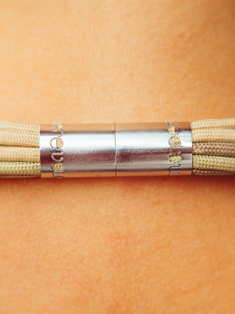 May include: Close-up of a necklace with a silver clasp and beige cords. The clasp is cylindrical with engraved text. The cords are tightly woven, displaying a gradient of beige and light green hues. The necklace is against a skin background.