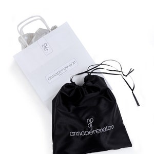May include: A white paper shopping bag and a black satin drawstring bag, both featuring the "annaperevalov" logo in a minimalist font. The bags are set against a white backdrop. The drawstring bag has a black ribbon.