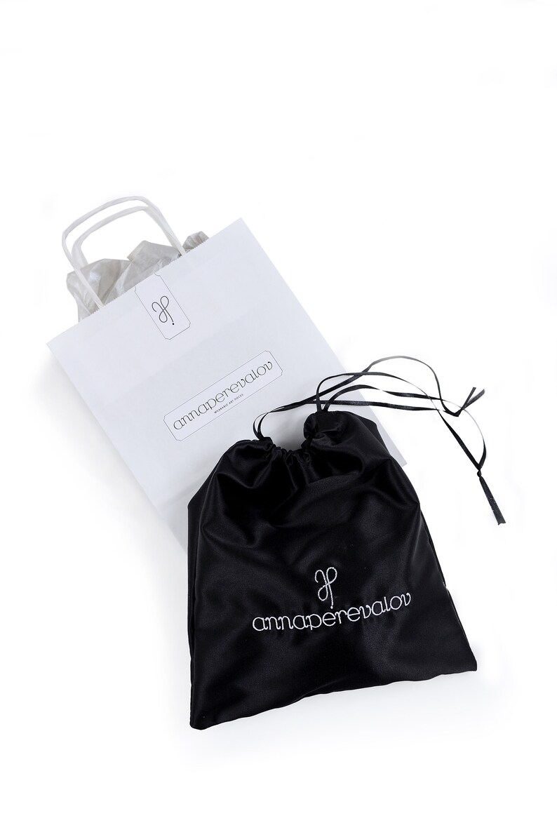 May include: A white paper shopping bag and a black satin drawstring bag, both featuring the "annaperevalov" logo in a minimalist font. The bags are set against a white backdrop. The drawstring bag has a black ribbon.