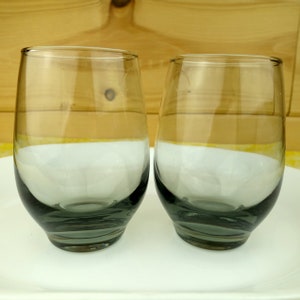 May include: Two clear glass tumblers with a dark gray tint. The glasses have a rounded bottom and a wide mouth.