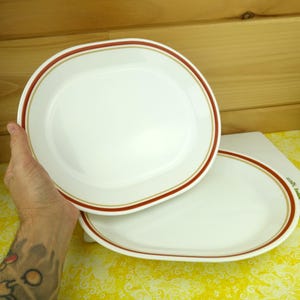 May include: Two white oval plates with a red and gold border. The plates are clean and appear to be made of ceramic or porcelain. The plates are on a yellow surface.