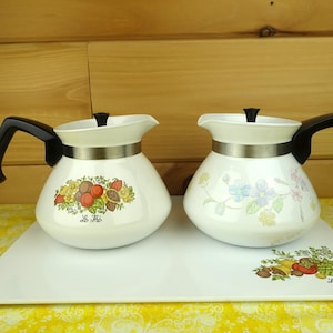 May include: Two white ceramic teapots with black handles and lids. One teapot features a fruit and vegetable design with the text "Le Thé". The other teapot has a floral design. Both teapots have a silver band.