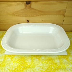May include: White ceramic baking dish with a slightly rounded rectangular shape and a decorative design around the rim. The dish is sitting on a yellow and white patterned surface.