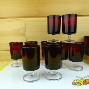 May include: A collection of vintage ruby red glasses. The glasses have a cylindrical bowl and a clear, circular base. The glasses are arranged on a white surface with a yellow and brown wooden background.