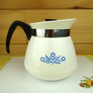 May include: A white teapot with a black handle and a black lid. The body of the teapot features a blue floral pattern consisting of three flowers and leaves. A silver-colored metal band encircles the upper portion of the teapot below the lid.