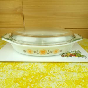 Puede incluir: Una fuente para horno ovalada con tapa de cristal transparente. La fuente blanca presenta una banda decorativa de motivos florales naranjas y marrones. La fuente está sobre una bandeja blanca con un diseño colorido de frutas y verduras.