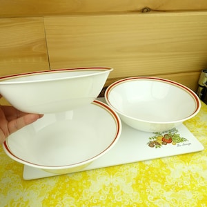 May include: Three white ceramic bowls with a red and tan trim. The bowls are on a white cutting board with a fruit and vegetable design. The cutting board is on a yellow patterned surface.