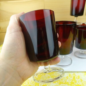 May include: A set of vintage ruby red wine glasses. The glasses have a rounded bowl and a clear stem and base. The glasses are held in a hand, with other glasses visible in the background. The glasses are likely made of glass.