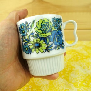 May include: White ceramic mug with a floral design featuring blue and lime green roses and other flowers. The mug has a unique handle and a ribbed base. The mug is held in a hand.