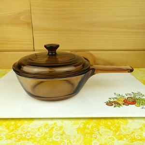 May include: A vintage amber-colored glass saucepan with a lid and a handle. The cookware is sitting on a white rectangular plate with a decorative vegetable design. The background is a light brown wooden surface.
