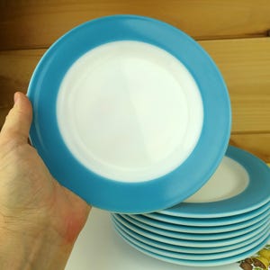 May include: A stack of round ceramic plates featuring a solid white center and a wide, bright turquoise blue rim. The plates have a smooth, glossy finish and a simple, circular design, presented as a set of matching dinnerware pieces.