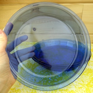 May include: A transparent blue Pyrex pie dish. The dish is round with a slightly raised rim and a clear, smooth surface. The Pyrex logo and other text are visible on the bottom of the dish. Made in the USA.