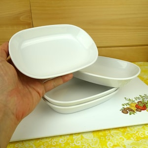 May include: A set of three white ceramic rectangular dishes with rounded corners and smooth, glossy surfaces. The dishes are stacked, with one dish held up separately to show its shallow depth and clean, minimalist white finish.