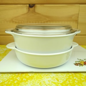 May include: A white glass baking dish with a glass lid. The dish is sitting on a white and yellow floral patterned surface.