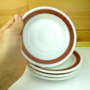 May include: A stack of white saucers with a red and gold decorative border. The saucers are round with a slightly concave center. The border features an ornate, repeating pattern. The saucers are stacked on a white surface.