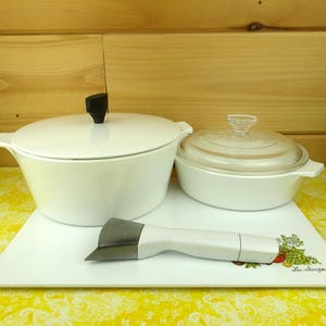 May include: White cookware set with lids, including a large pot with a black handle and a smaller dish with a glass lid. A white handled kitchen tool rests on a white rectangular serving tray.