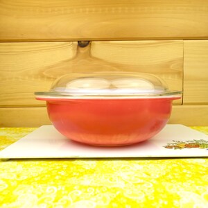 May include: A red glass casserole dish with a clear glass lid. The dish is sitting on a white surface with a floral pattern.