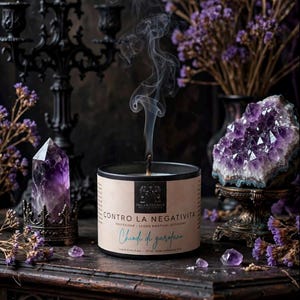 May include: A black candle in a cylindrical container with a beige label reading INCANTESIMUS CONTRO LA NEGATIVITA Chiodi di garofano. The candle emits a thin trail of smoke from its lit wick, surrounded by purple amethyst crystals and dried flowers.