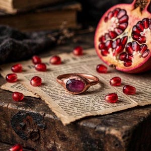 May include: A copper-toned ring featuring a dark red, oval-shaped gemstone set in a textured metal band. The ring rests on a piece of aged, printed paper surrounded by scattered, bright red pomegranate seeds and a halved pomegranate fruit.