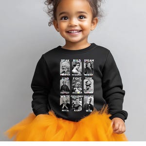May include: Black sweatshirt featuring portraits of historical figures with the words "Speak, Build, Dream, Lead, Fight, Think, Write, Challenge, Educate." The sweatshirt has a crew neck and long sleeves. The child is wearing an orange tutu.
