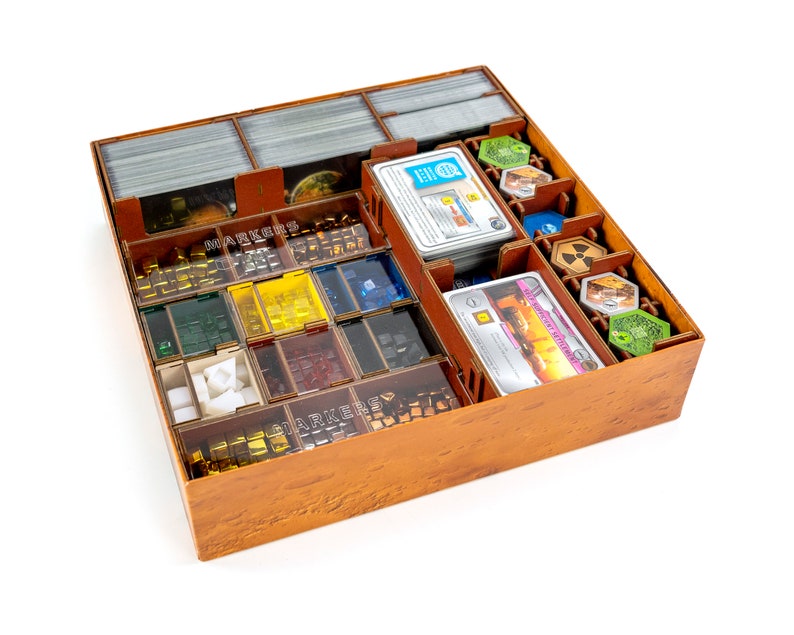 May include: A wooden game organiser with compartments for storing game pieces, cards, and tokens. The organiser has a brown wood finish and is filled with various game components, including small cubes, tiles, and cards.