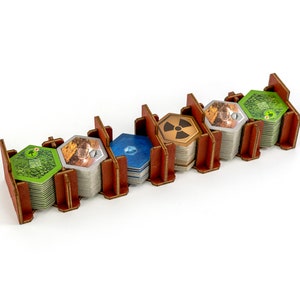 May include: A wooden organiser with five compartments for storing hexagonal game tiles. The compartments are filled with stacks of tiles in various colours, including green, brown, blue, and yellow. The tiles feature different symbols and designs.