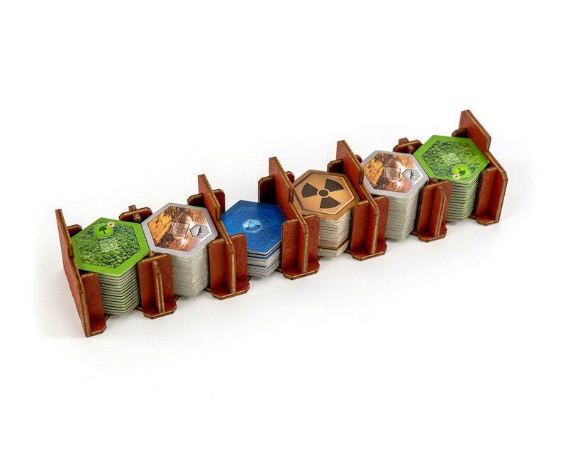 May include: A wooden organiser with five compartments for storing hexagonal game tiles. The compartments are filled with stacks of tiles in various colours, including green, brown, blue, and yellow. The tiles feature different symbols and designs.