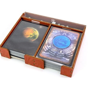May include: A wooden box with two compartments for storing cards. The box is brown with a clear plastic lid. The box is filled with cards featuring images of planets and a control panel.