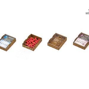 Pu&ograve; includere: Quattro scatole di legno con coperchi trasparenti, ciascuna contenente diversi componenti di gioco. Una scatola contiene cubi rossi, un'altra una carta con un'immagine di un ponte, e le altre due scatole contengono carte con testo.