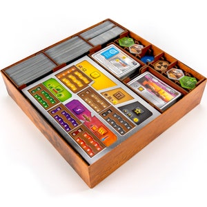 May include: A wooden box with a metal insert for organising game components. The insert has compartments for cards, tokens, and dice. The box is brown with a wood grain pattern. The insert is silver with black markings.