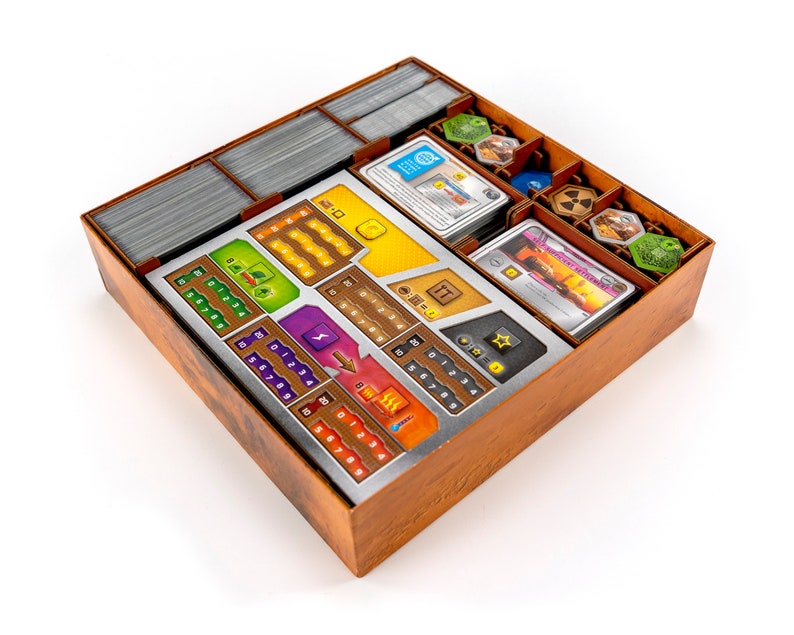 May include: A wooden box with a metal insert for organising game components. The insert has compartments for cards, tokens, and dice. The box is brown with a wood grain pattern. The insert is silver with black markings.