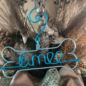 May include: A blue wire ornament shaped like a bone with the name "Emme" written on it. The ornament is hanging on a Christmas tree.
