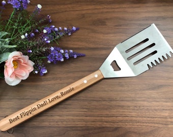May include: A wooden handled spatula with a metal blade. The blade has a serrated edge and a small hole in the center. The handle is engraved with the text "Best Flippin Dad! Love, Rossie".
