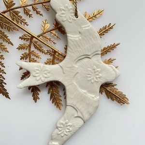 May include: White ceramic dove ornament with a floral design. The dove is hanging from a gold string.