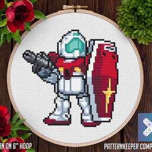 May include: A pixelated cross-stitch design of a robot warrior in a white, red, and teal color scheme, holding a gun and a red shield with a gold emblem. The design is set in a wooden embroidery hoop. Text reads "PATTERN ON 6" HOOP" and "PATTERN KEEPER COMPATIBLE."