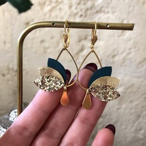 以下が含まれることがあります： フローラルデザインのゴールドトーンのドロップイヤリング。各イヤリングは、ティアドロップ型、ゴールドカラーのアクセント、ティール、ゴールドグリッター、ゴールドの重ねられた花びらを特徴としています。イヤリングはゴールドのスタンドに展示されています。